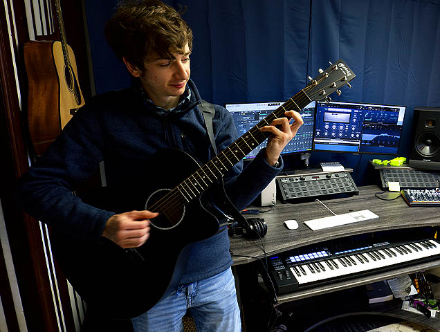 A student holding a guitar in front of a computer with music production equipment in the background.