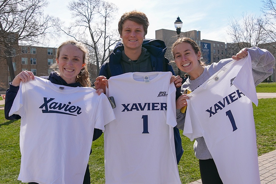 Three students pose together while holding out their new white Xavier t-shirts. They are in front of Kuhlman Hall.