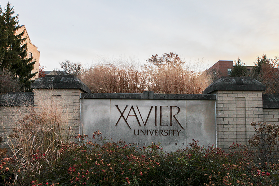 Stone sign marking the entrance to Xavier University's campus. Stone text reads 'Xavier University' and is flanked by brick columns. There are flowers and long grass planted around the sign.