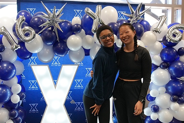 Two students post in front of a Xavier-themed photo backdrop with silver '16' balloons