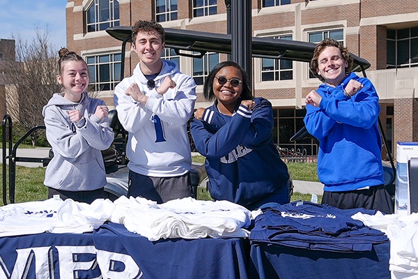 Four members of the X-Treme fans cross their arms to form an X. They are standing behind tables with stacks of Xavier t-shirts on them. 